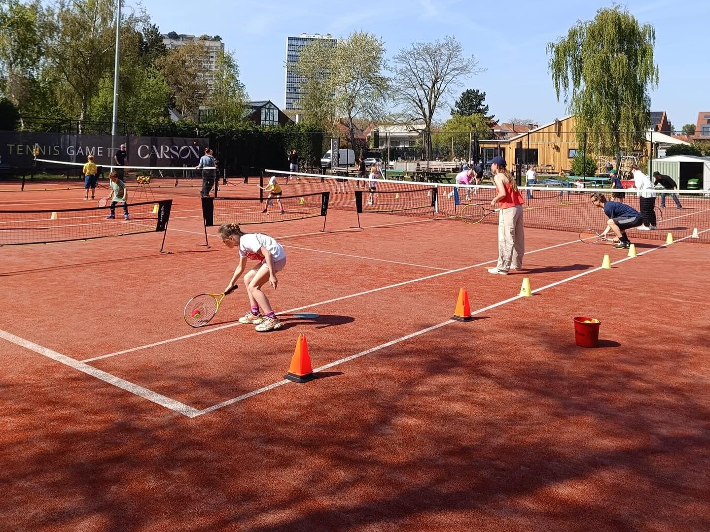 Kinderen in de tennisles op het gravel, met lage netten, pylonen en een ballenemmer